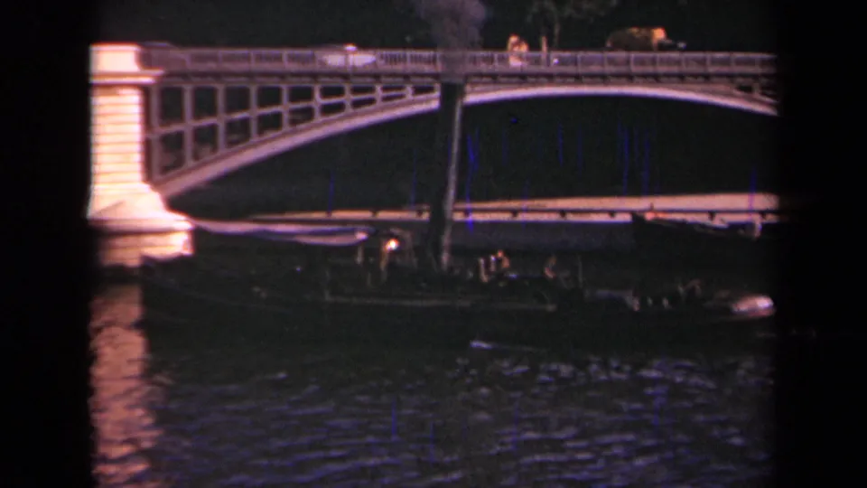 1939 Paris 8mm frame showing an illuminated arched bridge, river reflections, and a low passenger barge beneath it at night.