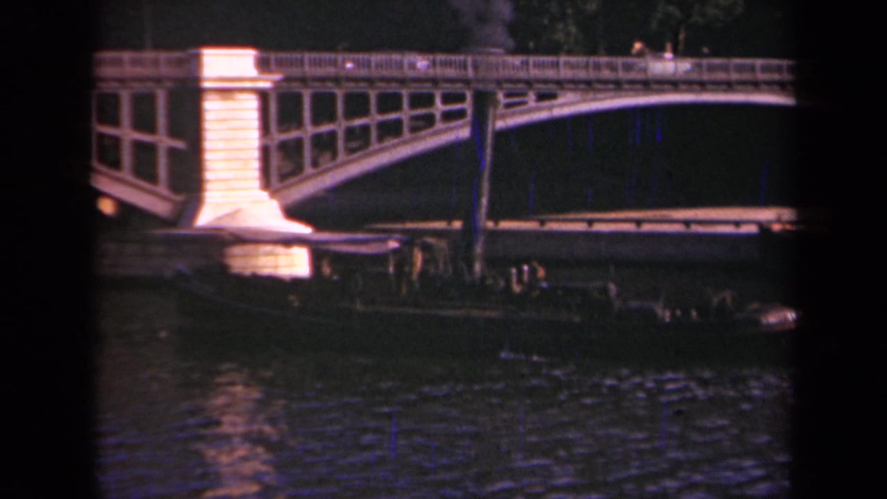 Night view in Paris showing the lit bridge pier and railing above a low river barge with passengers visible in silhouette.
