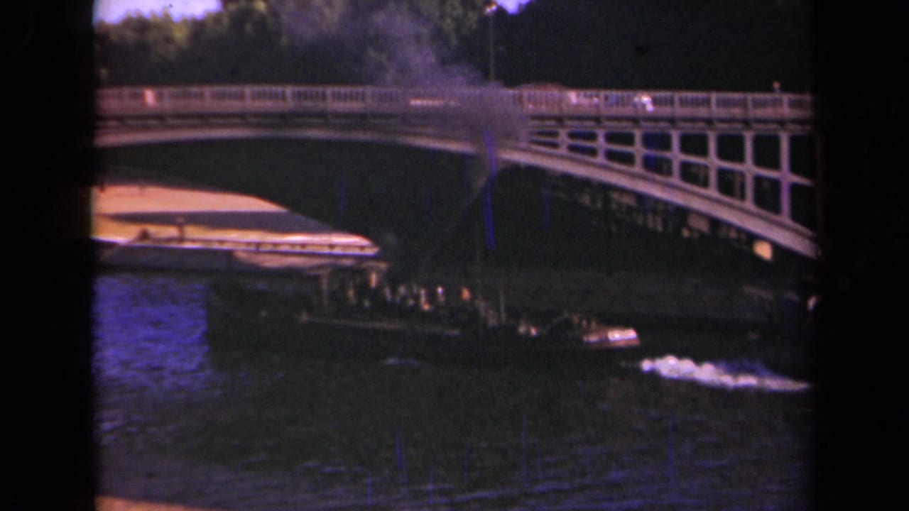 1939 Paris home-movie frame showing a small smoke-belching boat moving toward an arched bridge over dark water.