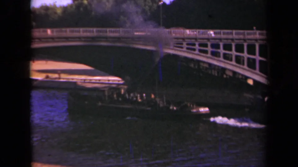 1939 Paris home-movie frame showing a small smoke-belching boat moving toward an arched bridge over dark water.