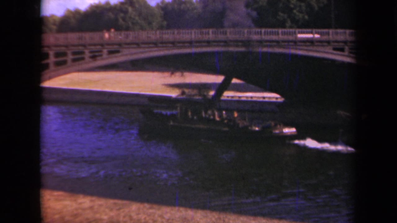 1939 Paris home-movie frame showing a boat approaching the shadow of an arched bridge while cars pass on the roadway above.