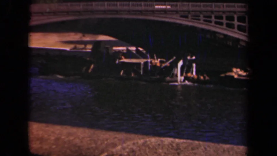 1939 Paris home-movie frame showing the boat entering the bridge shadow near riverside structures and small distant figures.
