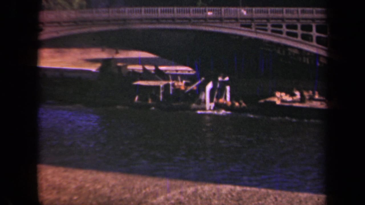 1939 Paris home-movie frame showing a low boat under an arched bridge with river water in the foreground.