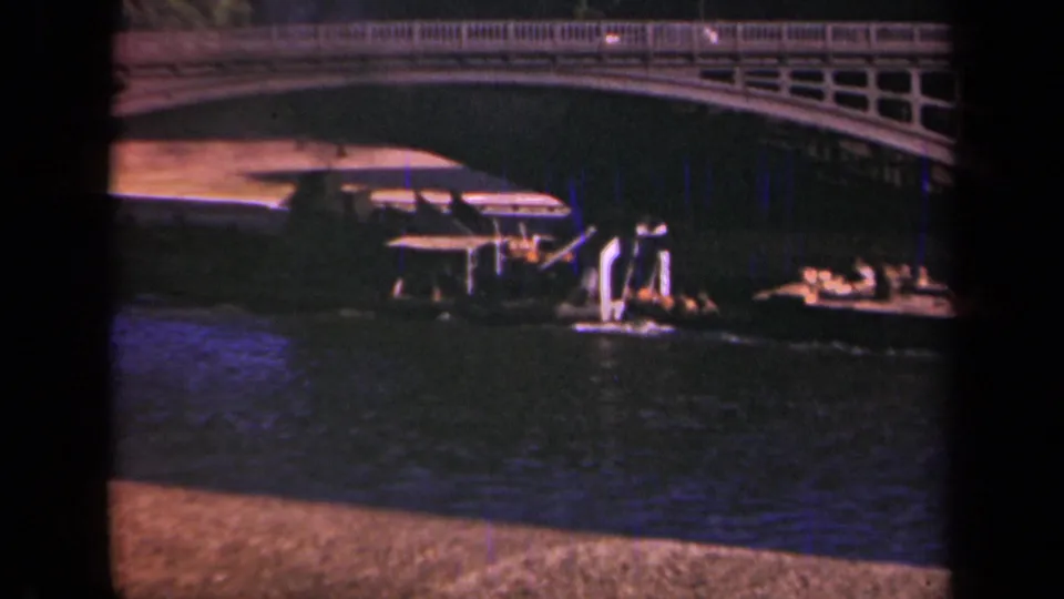 1939 Paris home-movie frame showing a low boat under an arched bridge with river water in the foreground.