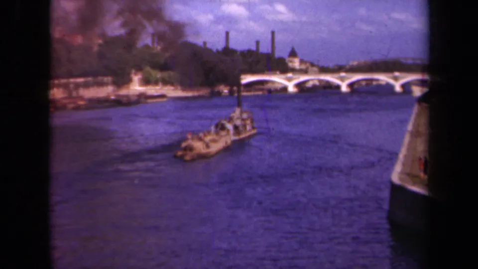1939 Paris home-movie frame showing a barge on the Seine, a multi-arched bridge, smokestacks, and dark smoke near the riverbank.