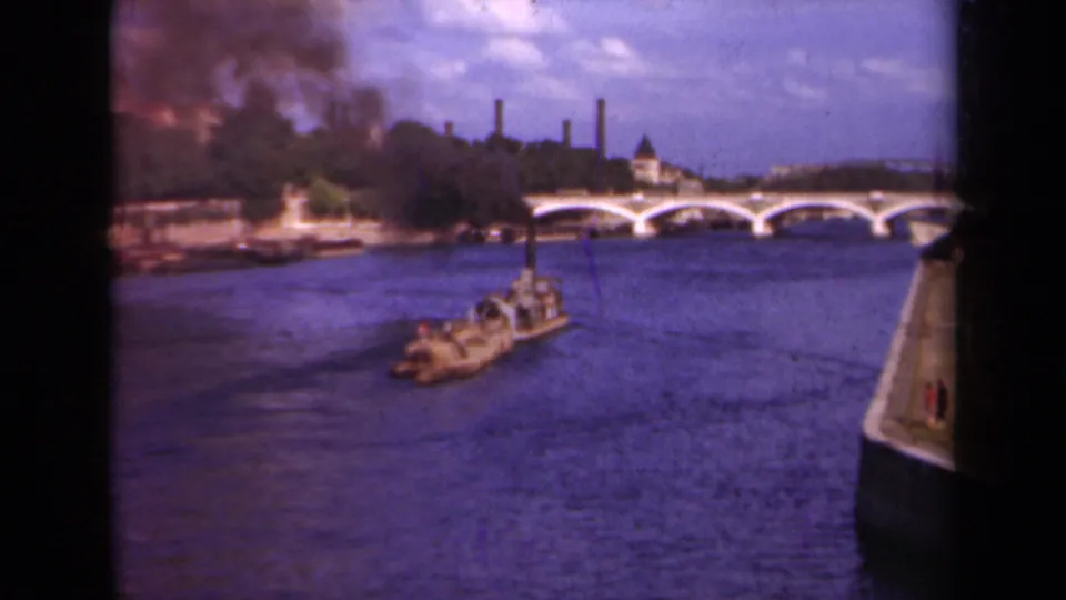 Soft 8mm frame of a barge on the Seine in Paris, with dark smoke rising beyond a tree-lined bank and bridge arches.
