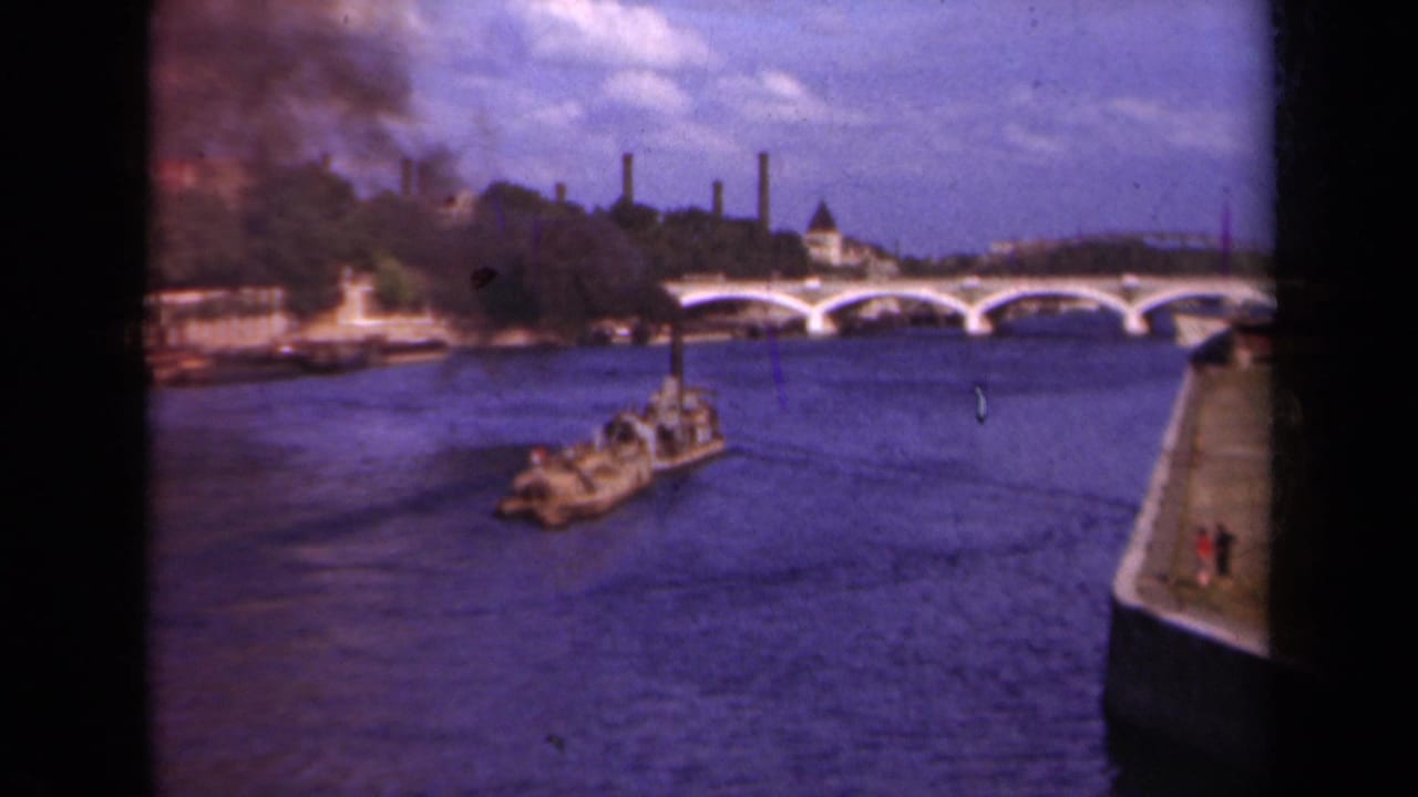 1939 Paris home-movie frame showing a small riverboat on the Seine with a bridge, quay, trees, and distant chimneys.