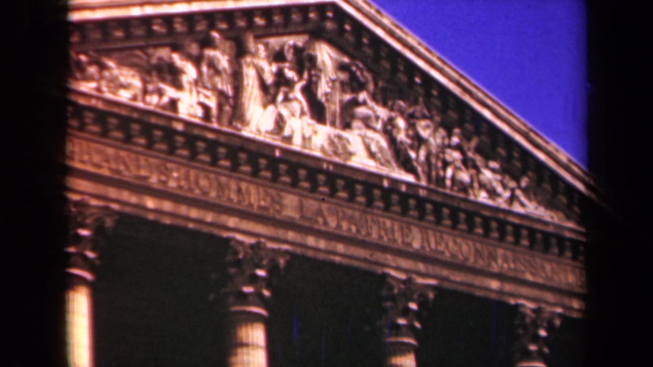 1939 Paris 8mm frame of a monumental classical facade, with sculpted figures, cornice detail, columns, and vivid blue sky.