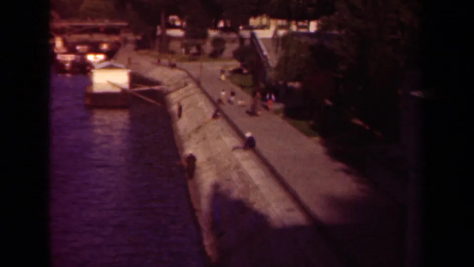 1939 Paris riverside view from above, showing a stone embankment, water, boats, trees, and distant people walking.