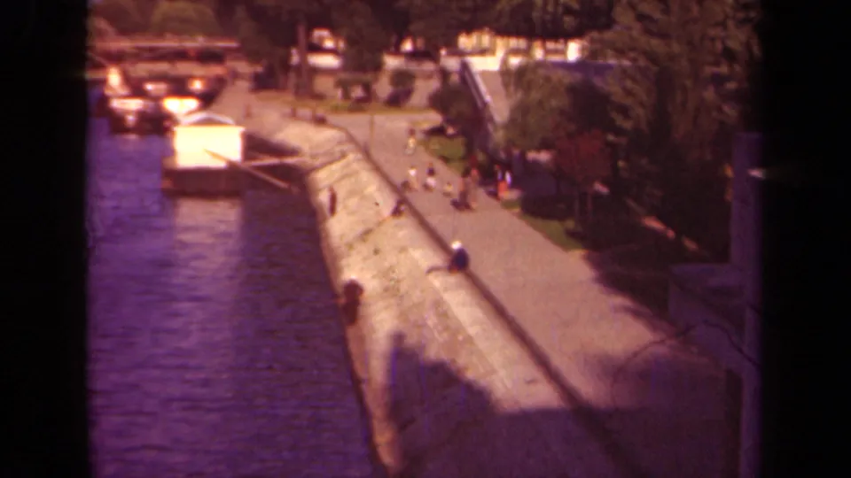 Elevated 8mm view of a Paris riverside quay with calm water, moored boats, trees, and distant pedestrians on the path.