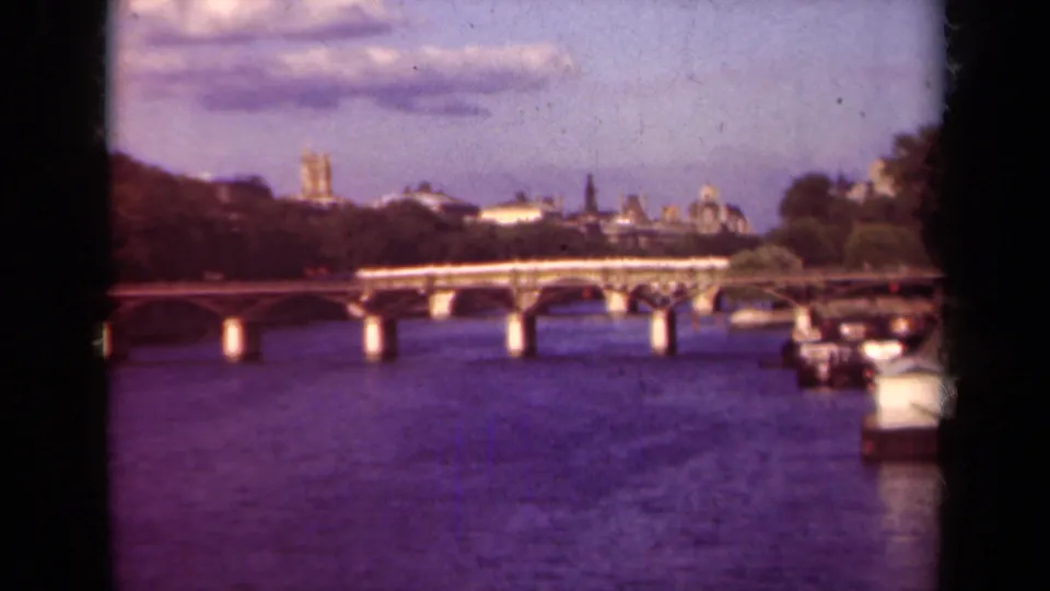 1939 Paris home-movie frame with the Seine, arched bridge, moored boats, and Notre-Dame towers visible on the skyline.