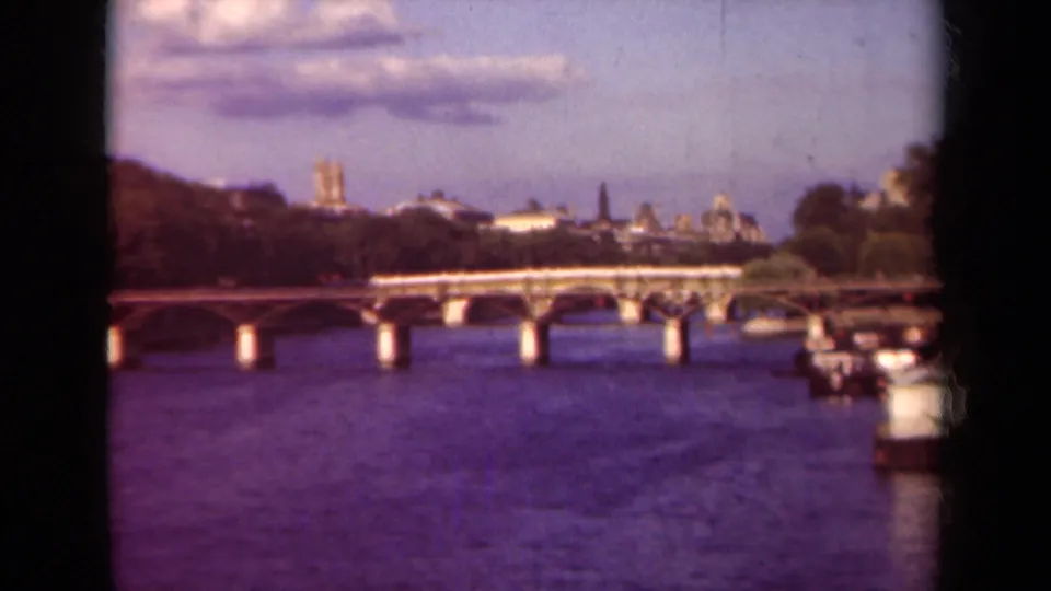 Soft 1939 Paris 8mm frame showing the Seine bridge, riverbank boats, trees, and distant Notre-Dame skyline under clouds.