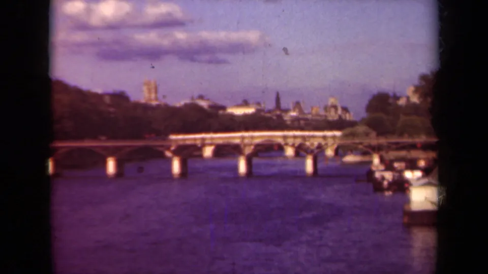 1939 Paris home-movie frame showing the Seine, a stone bridge, moored boats, and distant city landmarks under a blue sky.