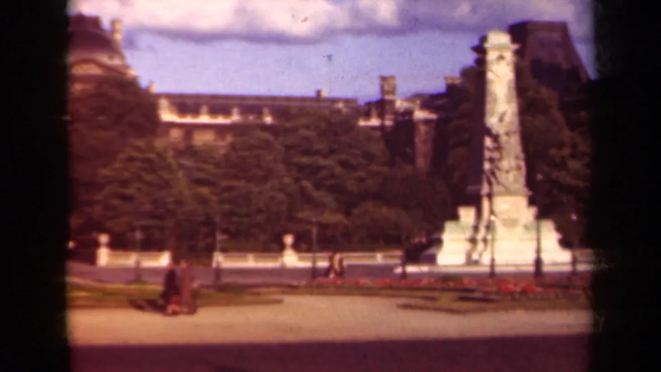 1939 Paris home-movie frame showing a tall stone monument, Louvre-side trees, paths, and distant pedestrians.