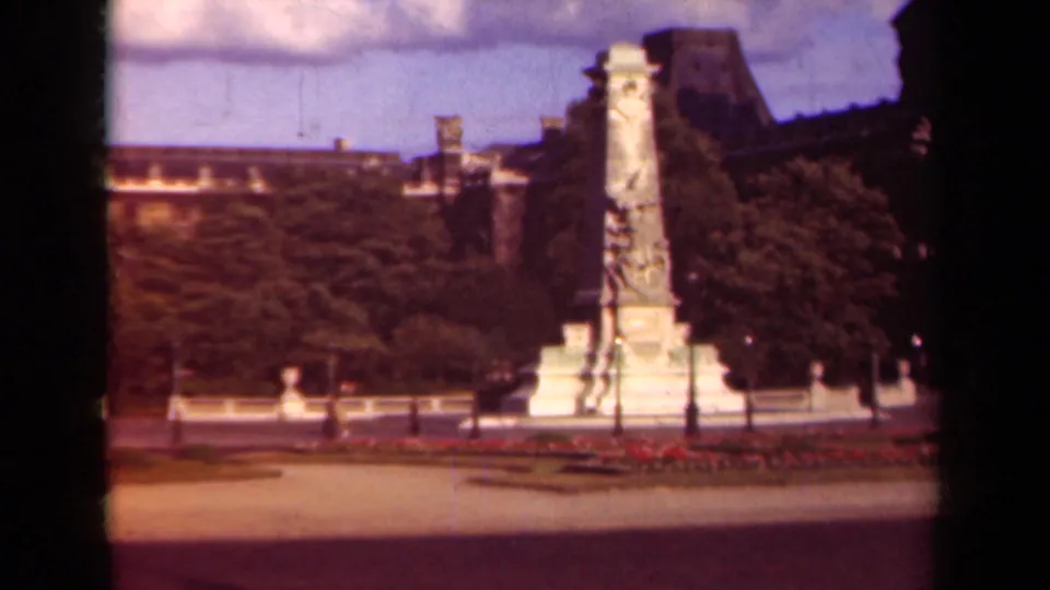 1939 Paris frame with a tall monument, flowerbeds, trees, and the Louvre-area plaza in soft 8mm color.