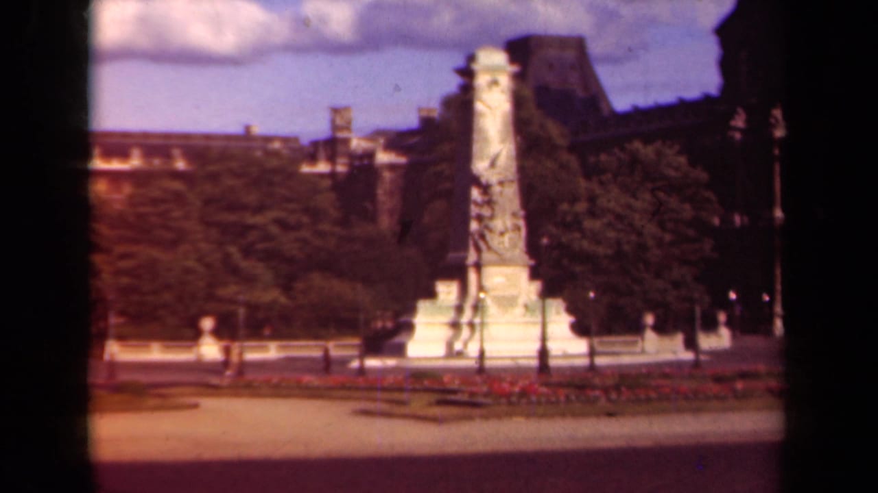 Paris square with a tall stone monument, trees, flowerbeds, lampposts, and distant pedestrians.