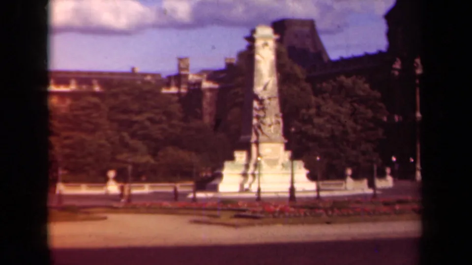 1939 Paris home-movie frame showing a formal square centered on a tall sculpted stone monument with gardens, trees, lampposts, and small pedestrians.