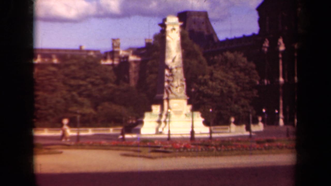 Wide view of the monument in a garden square with trees, lampposts, paths, and small figures.
