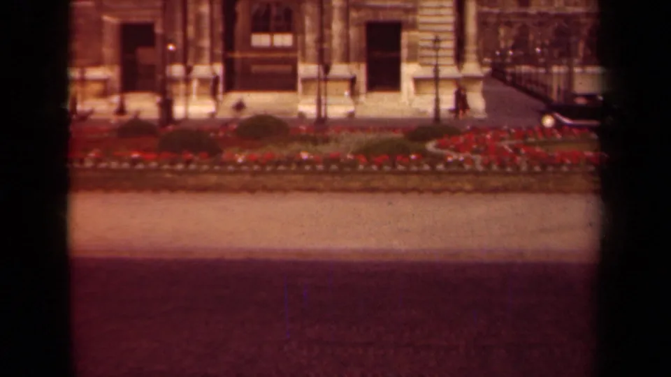 1939 Paris home-movie frame showing red flowerbeds, paths, lampposts, an ornate stone building facade, distant pedestrians, and a dark car at frame right.