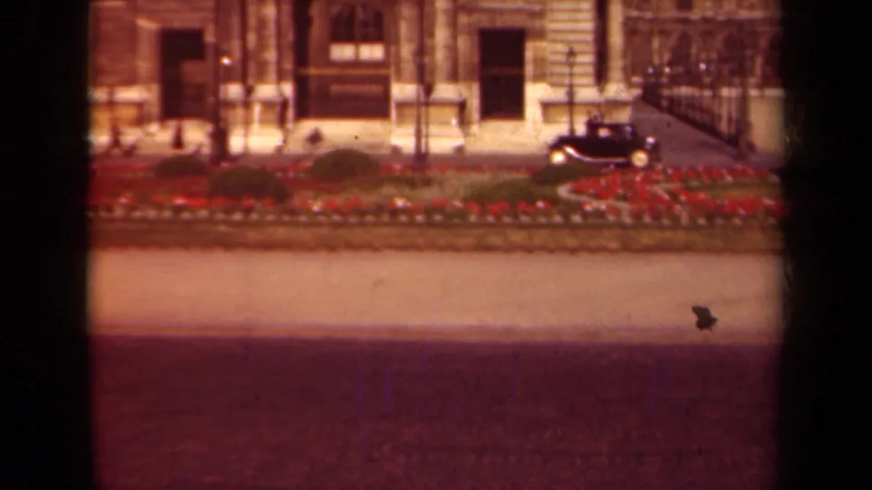 A 1939 Paris 8mm frame shows a dark rounded car moving past a stone facade, iron railings, and red median flowers.