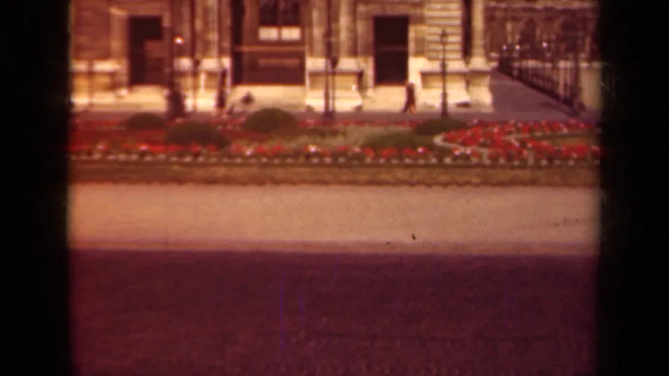 The later frame holds on the Paris streetscape with pedestrians, red median flowers, iron railings, and the formal stone building facade.