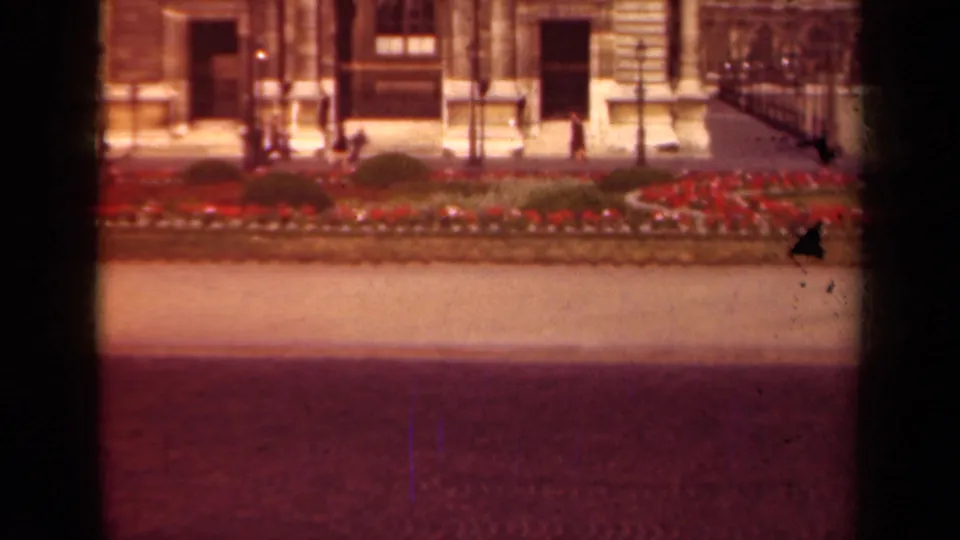 1939 Paris home-movie frame showing a formal stone facade, red flower beds, pavement, and small pedestrians in the distance.