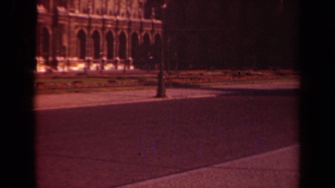 Stone arcade, lamppost, flower beds, trees, and open pavement viewed through dark film edges.