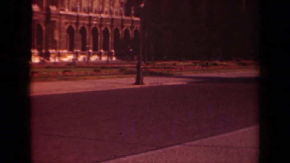 1939 Paris frame with a grand arched facade, lamppost, landscaped beds, and broad pavement under warm faded color.