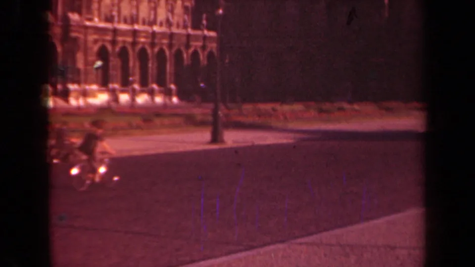 1939 Paris home-movie frame showing a bicyclist with a glowing front light passing a lamppost near an ornate historic building.