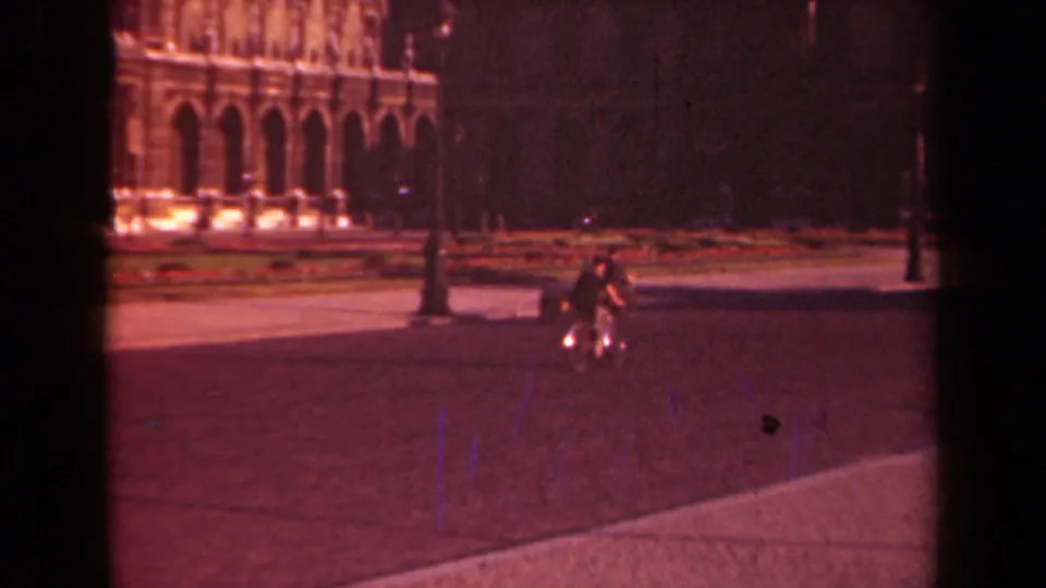 1939 Paris home-movie frame showing the bicyclist approaching through the square, with ornate arches and a lamppost in the background.