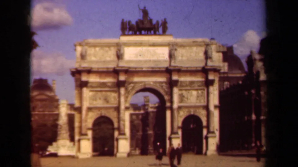 Paris landmark view from 1939 home-movie footage, showing a triumphal arch with statues, relief panels, pedestrians, and surrounding buildings.