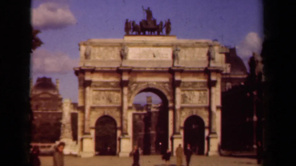Soft 8mm view of a Paris triumphal arch, with a pale column at left and pedestrians near the monument base.