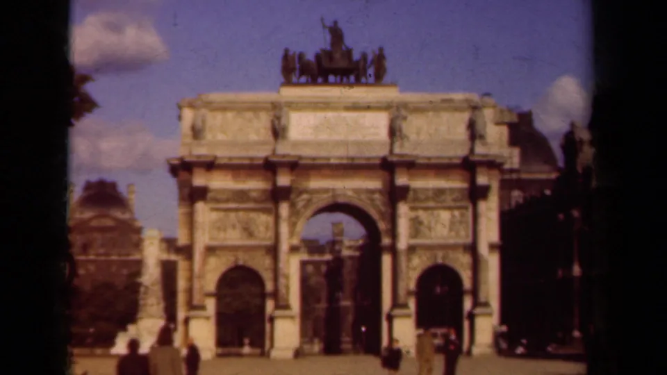 Soft 8mm frame of a Paris triumphal arch, with visitors crossing the foreground under a bright sky.