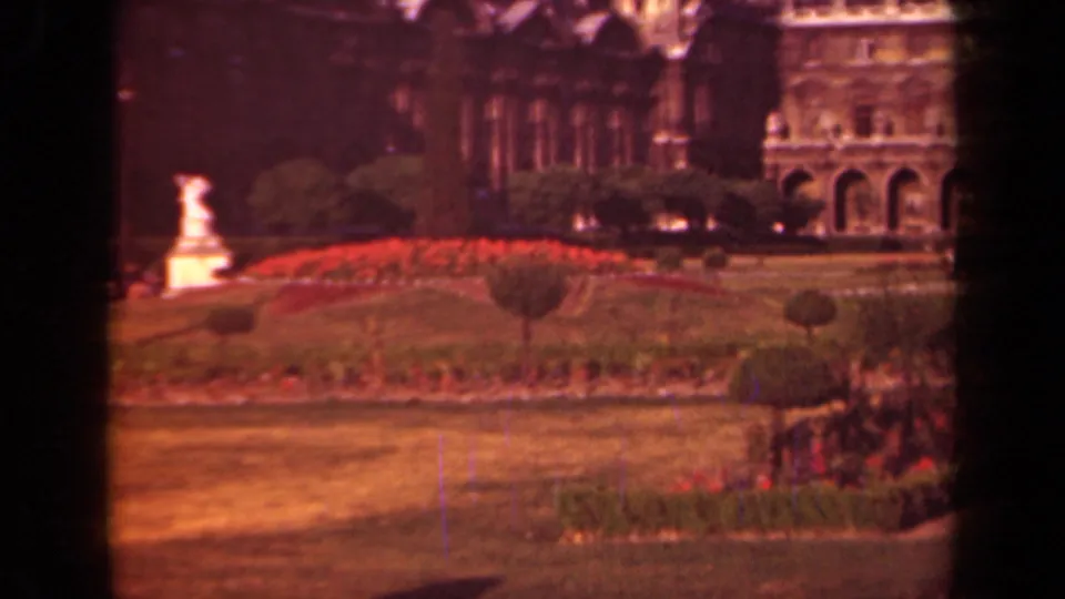 1939 Paris frame showing the formal garden from a fixed visitor viewpoint, with red flowerbeds, a white statue, clipped lawn, shrubs, and ornate stone building details.