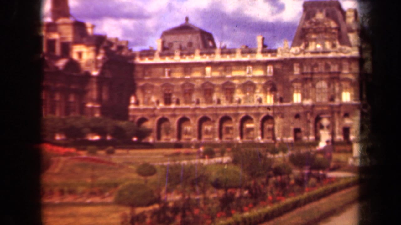 The Louvre exterior appears beyond formal lawns, red flowerbeds, hedges, and a cloudy sky in 1939 Paris footage.