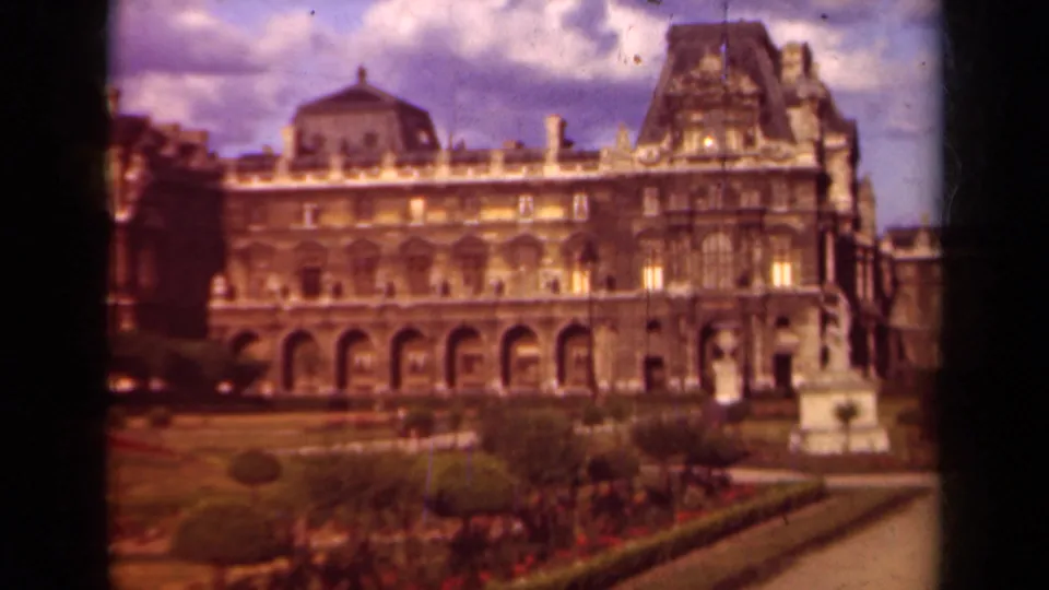 The Louvre facade stands behind formal gardens, hedges, red plantings, and a statue in this 1939 Paris home-movie frame.