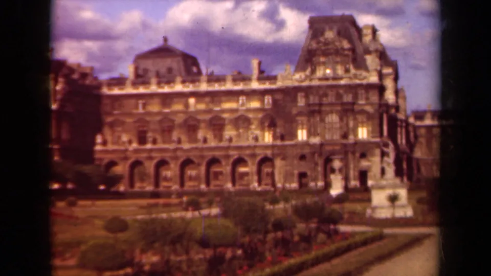 1939 Paris 8mm view of the Louvre palace facade, arched ground level, formal garden beds, and a partly cloudy sky.