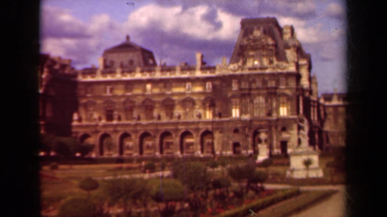 1939 Paris color 8mm frame showing the Louvre exterior across clipped garden beds, with arched bays and rooflines visible.