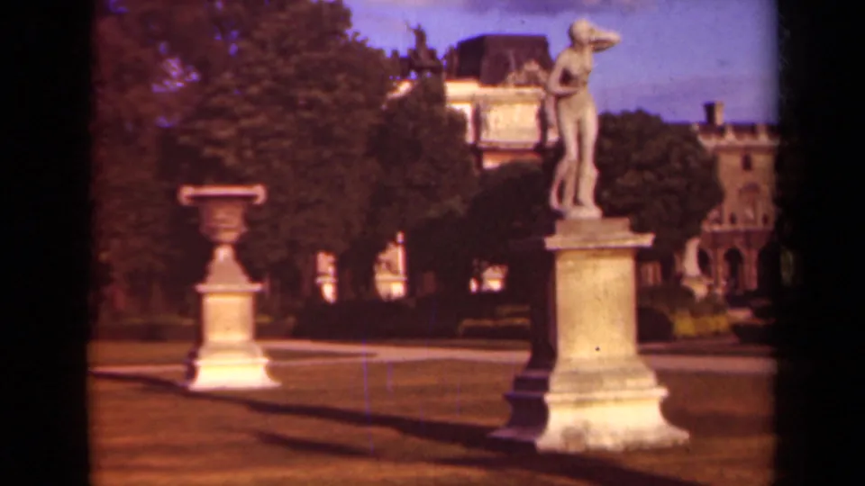 1939 Paris 8mm frame of a classical statue on a pedestal in the Louvre gardens, with trees, paths, urns, and palace rooflines visible.