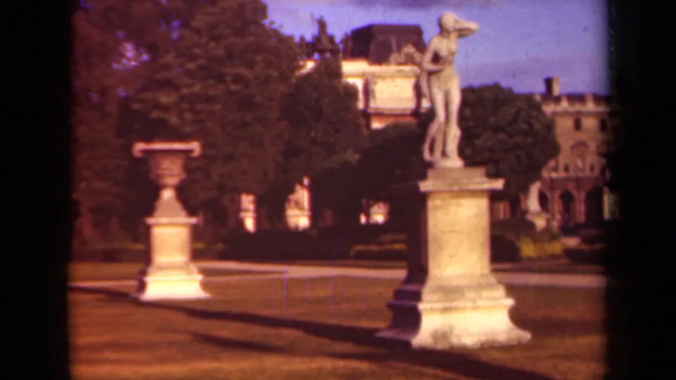 1939 Paris home-movie frame showing a classical statue, urn, lawn, trees, and historic building backdrop.