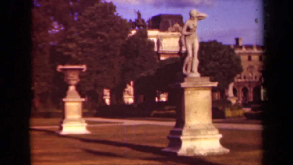 Warm 8mm Paris park view with a classical statue, urn, lawn, trees, and masonry building beyond.