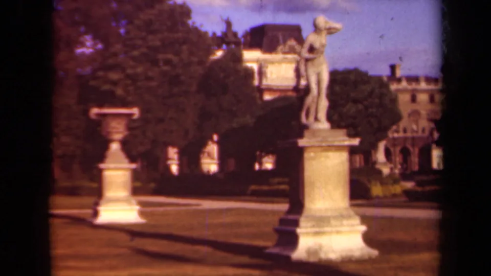 1939 Paris home-movie frame showing a classical garden statue on a pedestal, a stone urn, lawn, trees, and distant architecture.
