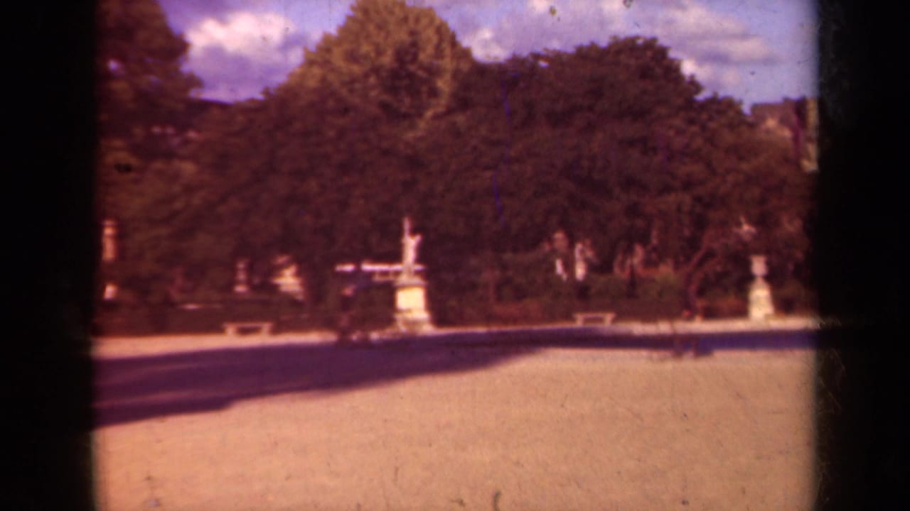 1939 Paris garden view with pale gravel path, classical statues on pedestals, dense trees, and a distant archway under clouds.
