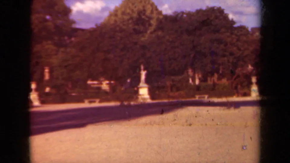 1939 Paris home-movie frame of a formal garden court with a statue on a pedestal, tree line, pale path, and visible film wear.