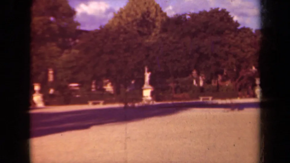1939 Paris garden frame showing a statue across a sunlit path, framed by dense trees and dark 8mm gate edges.