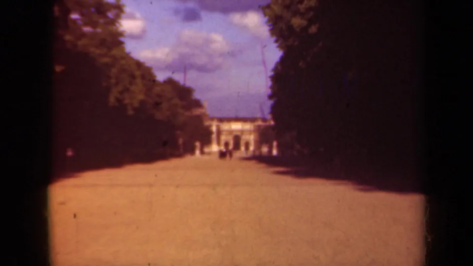 1939 Paris frame showing a formal walkway bordered by trees, with pedestrians and a pale classical building in the distance.