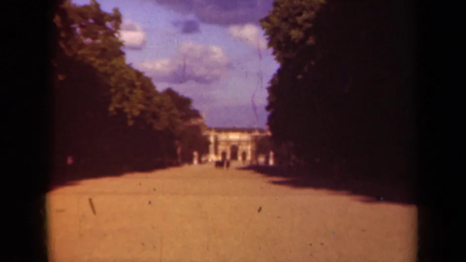 1939 Paris 8mm still of a sunlit tree-lined walkway approaching a distant formal building with indistinct people nearby.