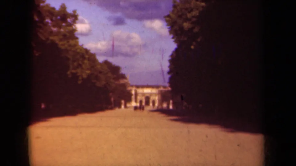 1939 Paris park view with a sunlit avenue, shaded tree rows, small distant visitors, and a classical building ahead.
