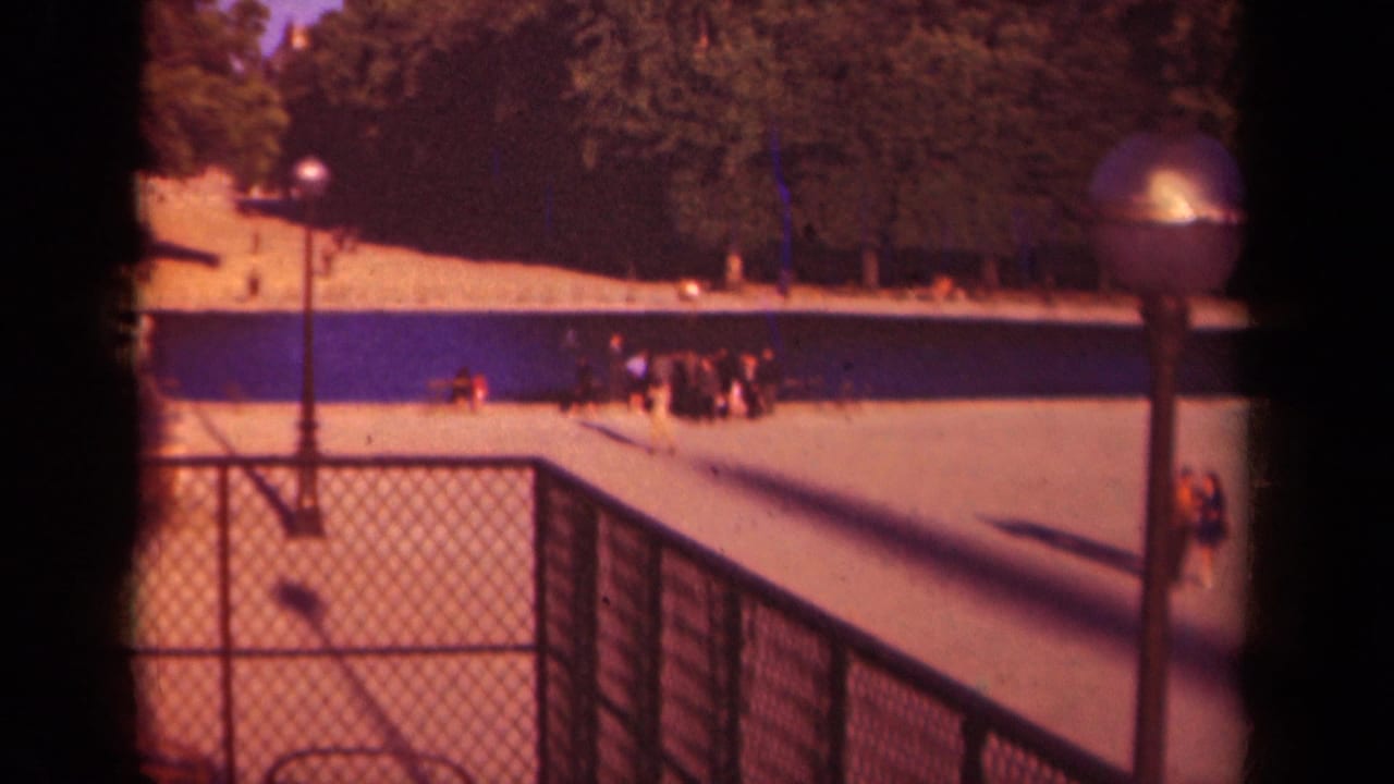Distant people in a Paris park setting stand and walk near a paved promenade, blue water feature, foreground fence, and tree-lined background.