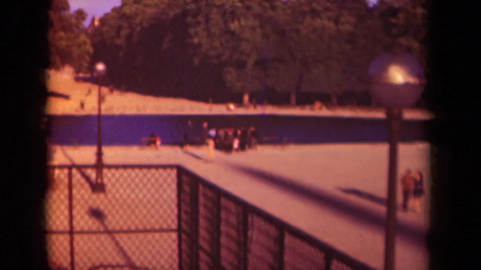 1939 Paris park footage shows distant visitors on a paved walkway, with a chain-link fence in front, lampposts, trees, and warm faded film color.